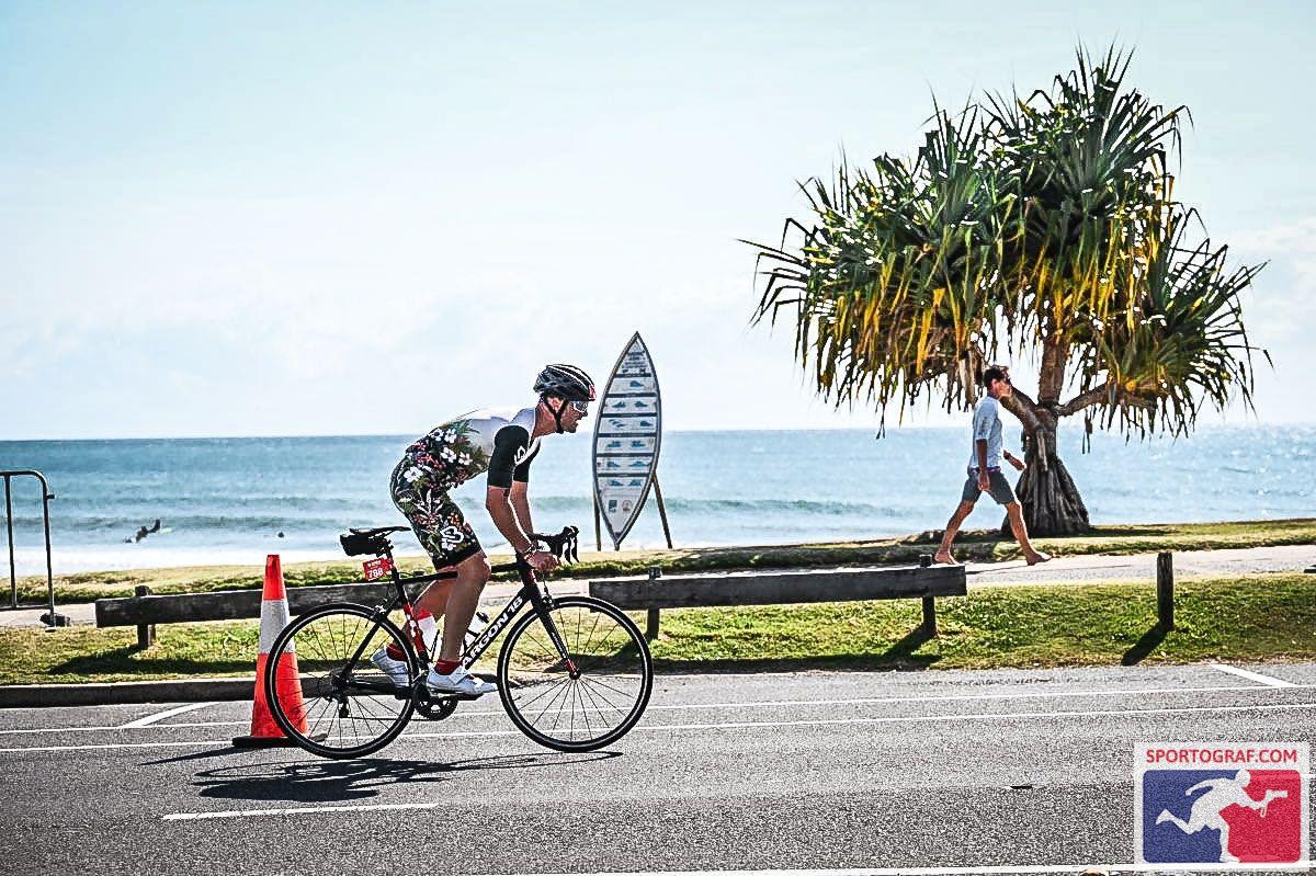 A man riding a bicycle wearing a helmet and protective glasses. A beach is captured in the background.