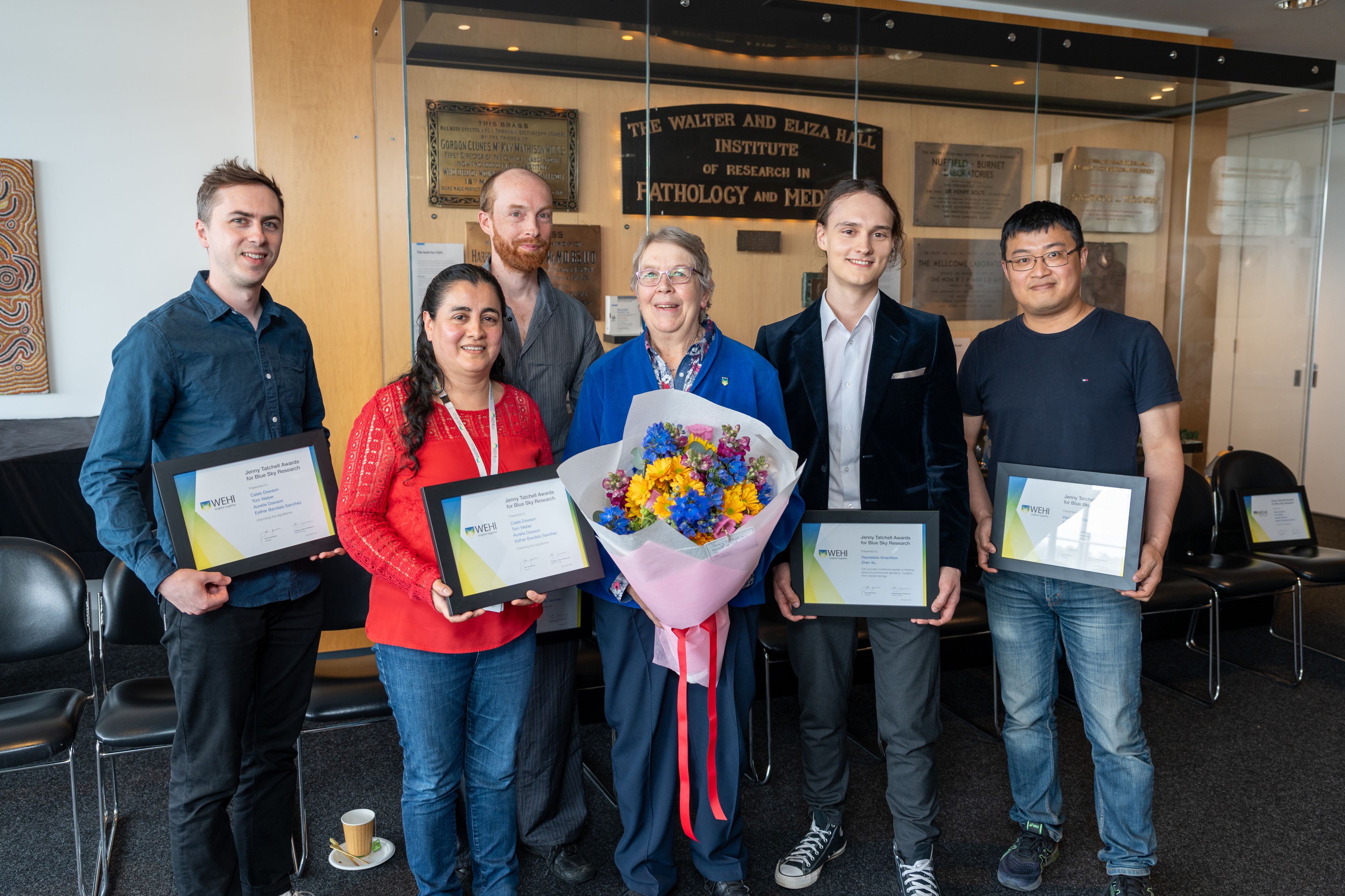 A woman and two men standing on the left each holding a framed certificate, a woman in the middle holding a bouquet of flowers and two men on the right holding a framed certificate.