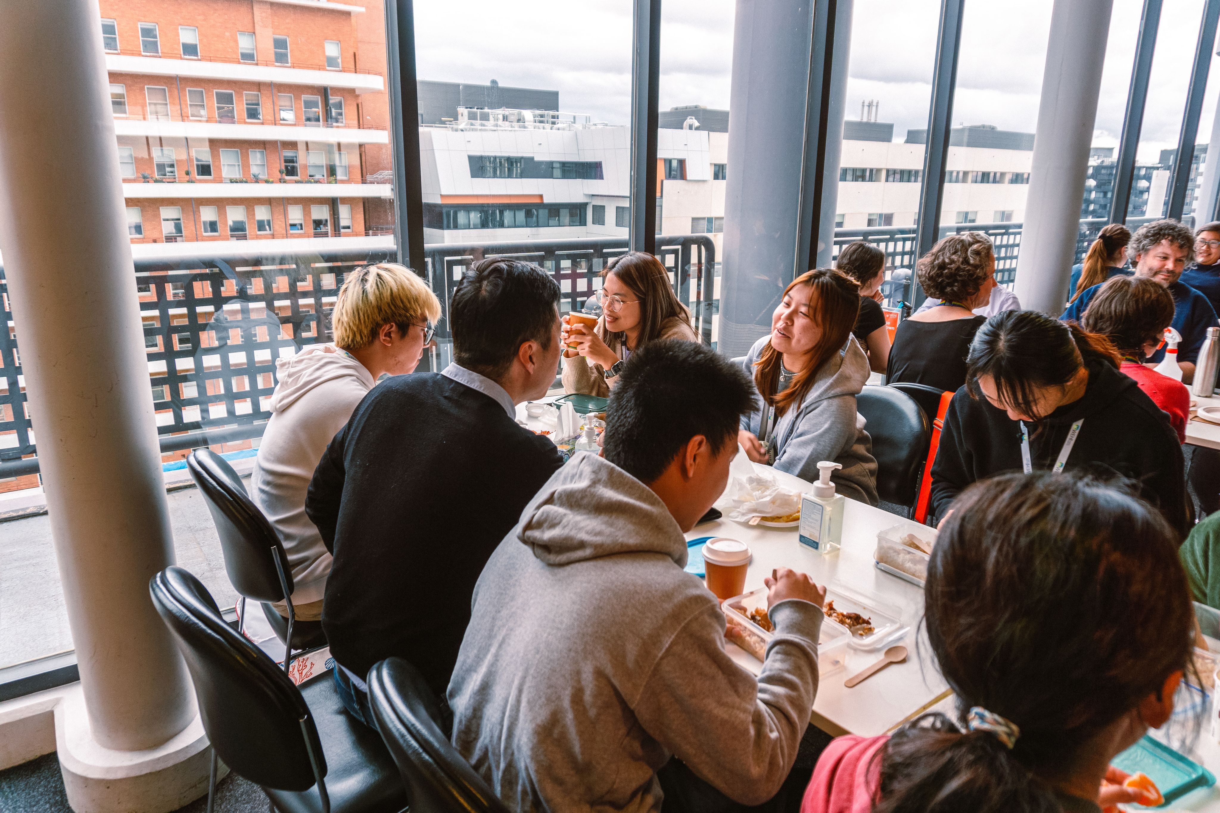 A group of young men and woman sitting at a table. The individuals are captured talking to one another whilst eating their lunch.
