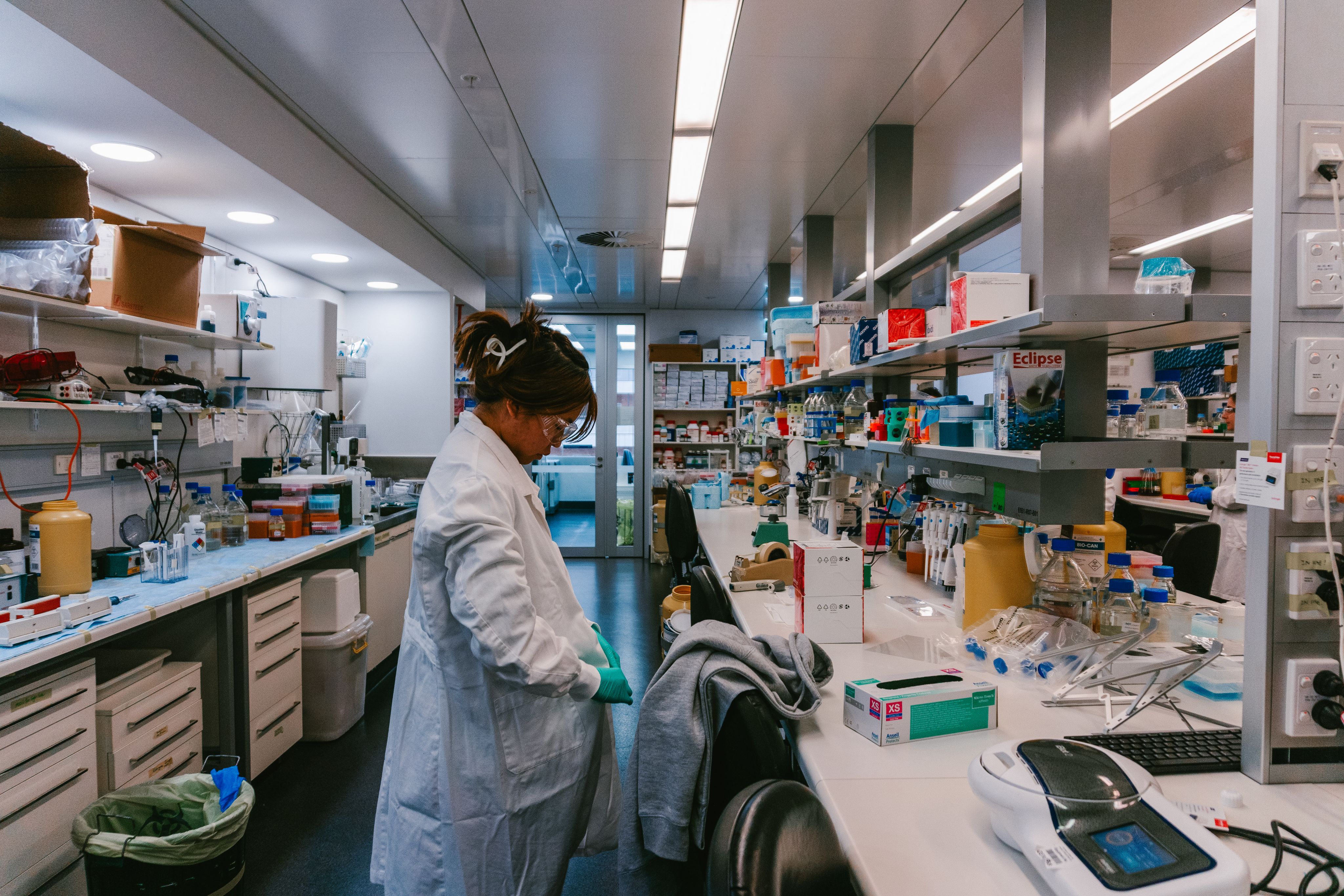 A woman standing in lab captured wearing protective glasses and buttoning her lab coat.
