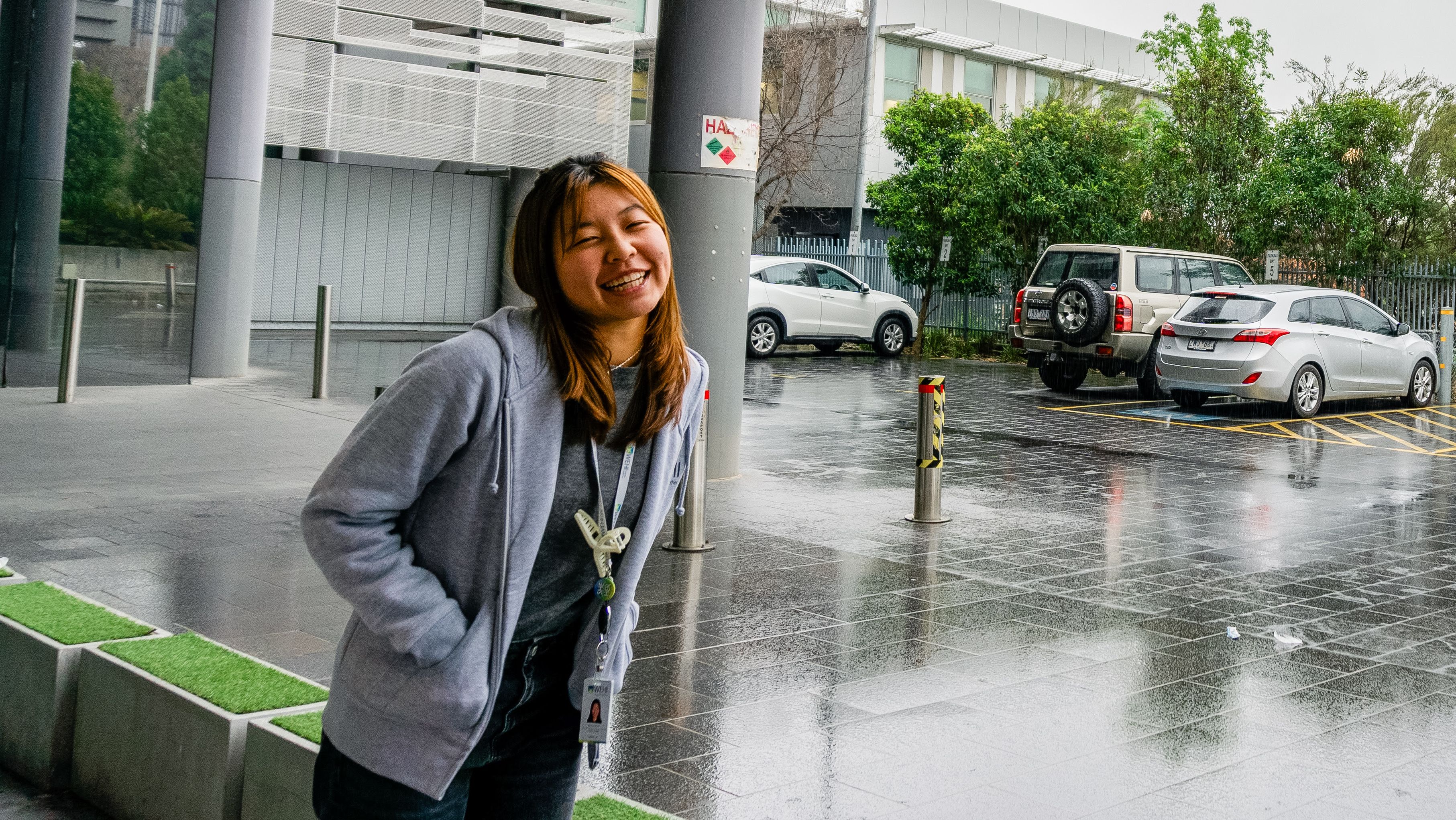 A young woman wearing a grey zip up jumper, smiling for a photo outside a building. 