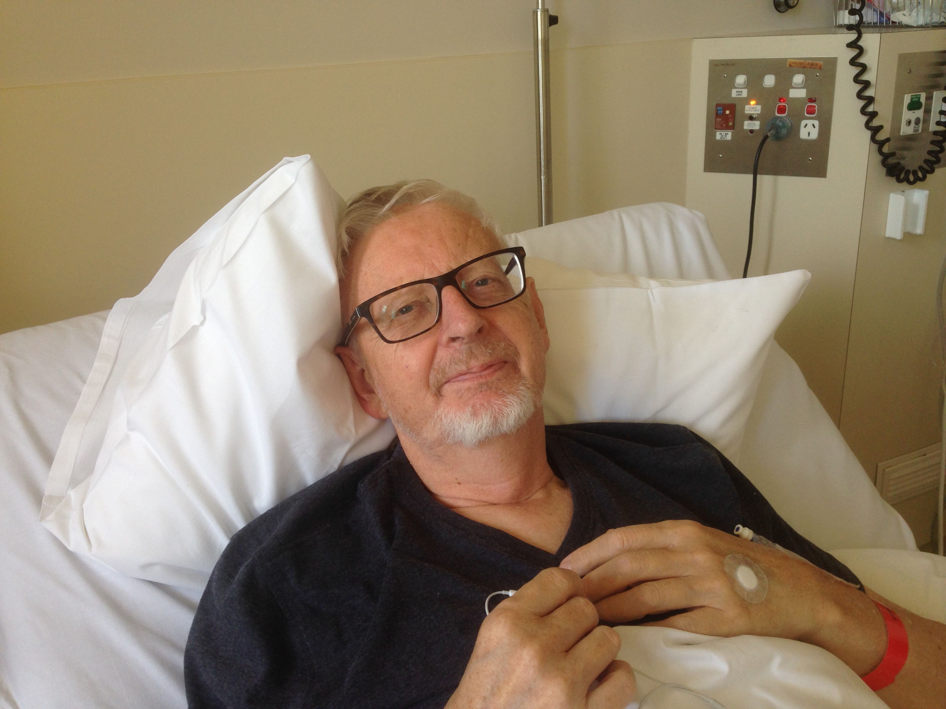 A man is lying in a hospital bed smiling directly towards the camera. The man is wearing glasses and a black t-shirt.