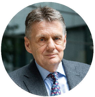 Circular cropped image of a man looking directly at the camera wearing a grey suit. There is bright natural lighting as the man is outside.