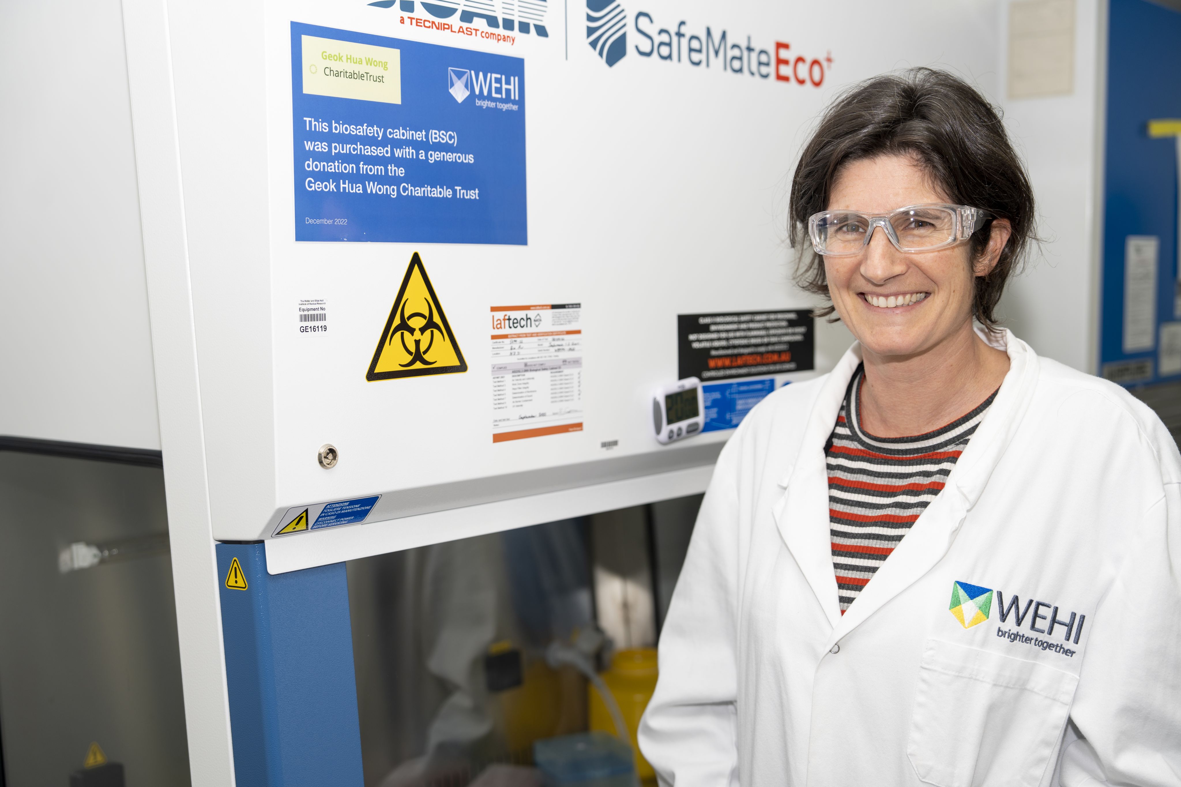 A woman positioned to the right of the image is smiling directly at the camera. The woman is wearing lab safety glasses and a lab coat.