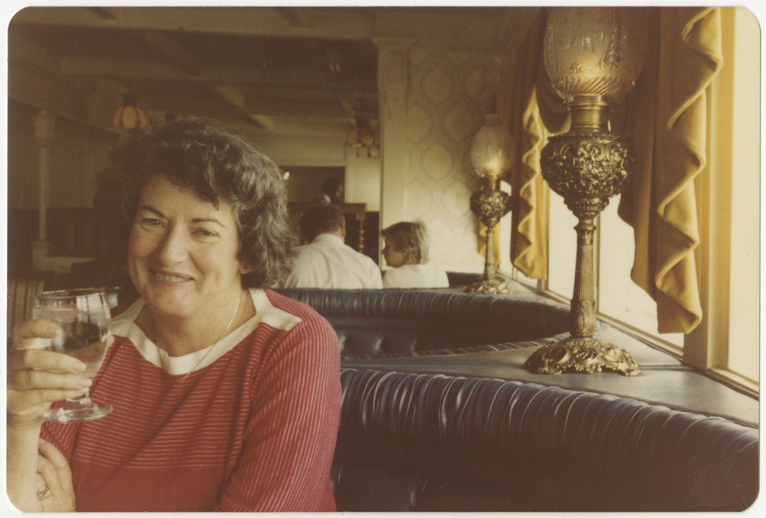 A woman wearing a red top is sitting at a restaurant booth holding a wine glass whilst smiling at the camera. 